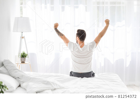 Relaxed young man waking up in bed and stretching his arms, facing the window, back view, free space 81254372