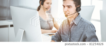 Casual dressed young man using headset and computer while talking with customers online in sunny office. Call center, business concept Casual dressed young man using headset and computer while talking with customers online in sunny office. Call center, business concept 81254826