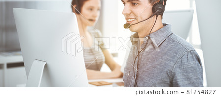 Casual dressed young man using headset and computer while talking with customers online in sunny office. Call center, business concept Casual dressed young man using headset and computer while talking with customers online in sunny office. Call center, business concept 81254827