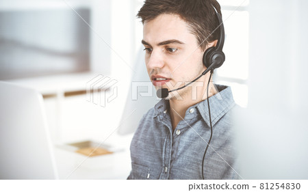 Casual dressed young man using headset and computer while talking with customers online in sunny office. Call center, business concept 81254830