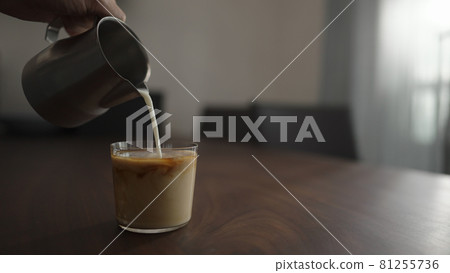pour milk into cold brew coffee over ice ball in tumbler glass on walnut table 81255736