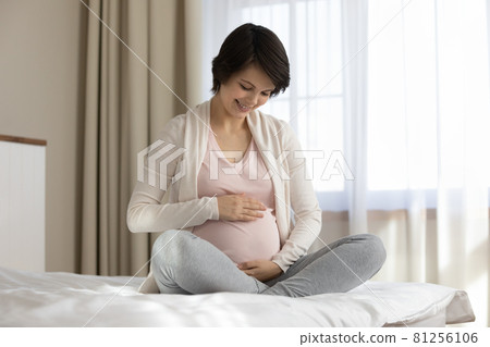 Happy young expectant mom sitting on bed, caressing big belly Happy young expectant mom sitting on bed, caressing big belly 81256106