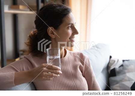 Woman sit indoor holding glass of water looks into distance 81256249