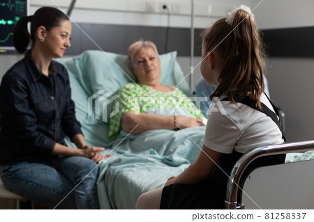 Selective focus on young granddother visiting sick retired grandmother in hospital ward 81258337
