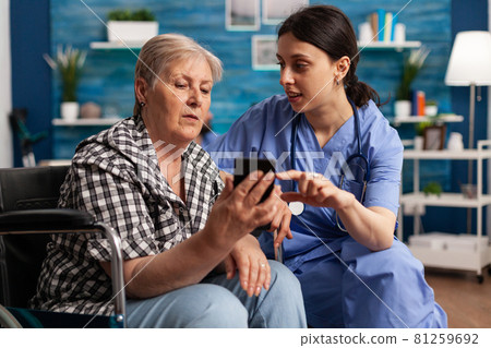 Nurse helping retired senior woman in wheelchair to use smartphone during social service 81259692