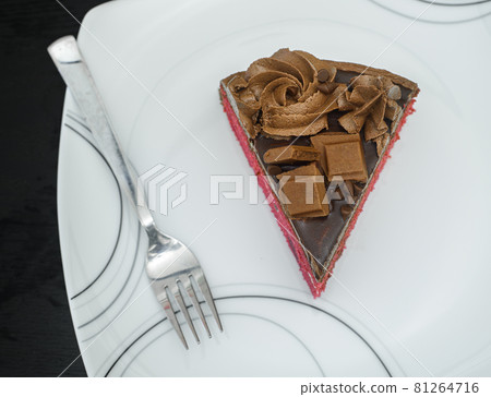 Chocolate cake slice and a fork in a white ceramic plate. Icing flowers and chocolate pieces as toppings. Delicious Icing Birthday cake design overhead close up view. Chocolate cake slice and a fork in a white ceramic plate. Icing flowers and chocolate pieces as toppings. Delicious Icing Birthday cake design overhead close up view. 81264716