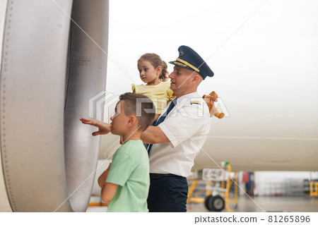 Pilot in uniform showing parts of airplane to two little kids who came to excursion at the aircraft hangar 81265896