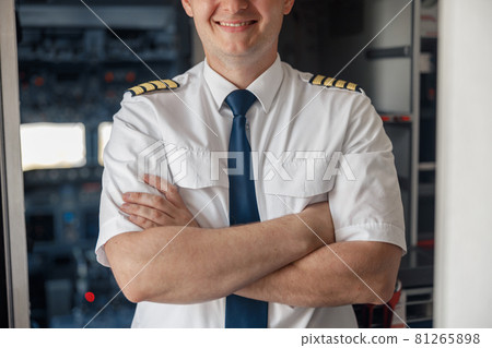 Cropped portrait of male pilot in uniform keeping arms crossed and smiling while posing, standing inside of the airplane Cropped portrait of male pilot in uniform keeping arms crossed and smiling while posing, standing inside of the airplane 81265898