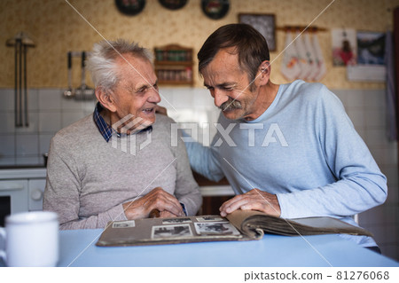 Portrait of man with elderly father sitting at the table indoors at home, looking at photo album. Portrait of man with elderly father sitting at the table indoors at home, looking at photo album. 81276068