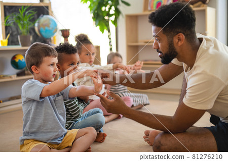 Group of small nursery school children with man teacher sitting on floor indoors in classroom, doing exercise. Group of small nursery school children with man teacher sitting on floor indoors in classroom, doing exercise. 81276210