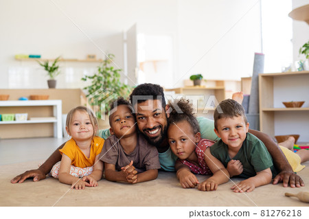 Group of small nursery school children with man teacher on floor indoors in classroom, looking at camera. Group of small nursery school children with man teacher on floor indoors in classroom, looking at camera. 81276218