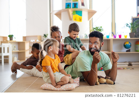 Group of small nursery school children with man teacher sitting on floor indoors in classroom, playing. 81276221
