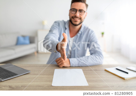 Job interview concept. Positive millennial man stretching hand for handshake, sitting at workplace, selective focus Job interview concept. Positive millennial man stretching hand for handshake, sitting at workplace, selective focus 81280905