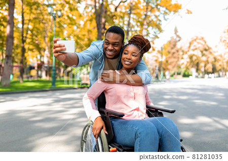 Cheerful black guy hugging his disabled girlfriend in wheelchair, making selfie on smartphone at autumn park Cheerful black guy hugging his disabled girlfriend in wheelchair, making selfie on smartphone at autumn park 81281035