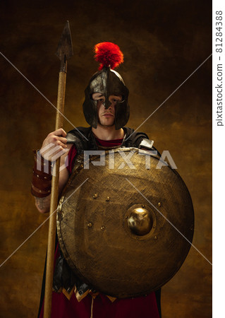 Portrait of medieval person, brutal man, warrior or knight in war equipment isolated on vintage dark background. Comparison of eras, art, history, retro style concept Portrait of medieval person, brutal man, warrior or knight in war equipment isolated on vintage dark background. Comparison of eras, art, history, retro style concept 81284388