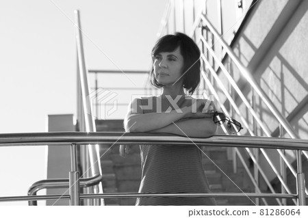 A brunette in a striped dress smiling stands on the stairs leaning her elbows on the railing and holding glasses in her hands 81286064