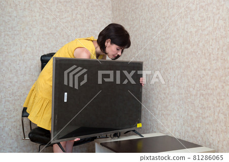 A brunette woman in the apartment installs a new large LCD TV on the table A brunette woman in the apartment installs a new large LCD TV on the table 81286065