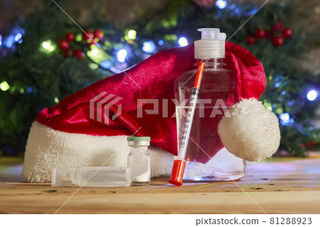 Covid-19 and Christmas holidays. Antiseptic hand sanitizer bottle, syringe and medicines on wood. Santa cap, coronavirus vaccines and syringe over Xmas tree 81288923