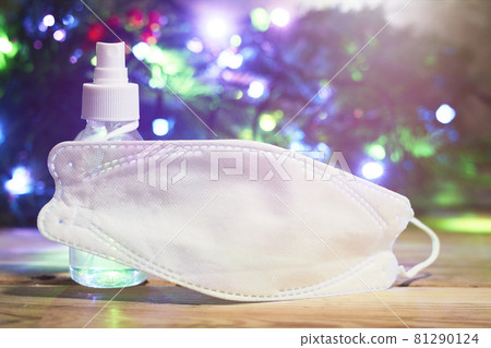 Covid-19 and Christmas holidays. Medical face mask and antiseptic gel over decorated with lights Christmas tree background 81290124