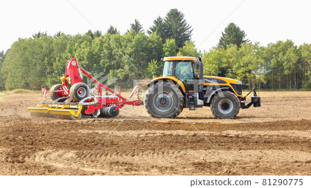 Yellow tractor pulls red sowing mechanism over dry field, trees in background. 81290775