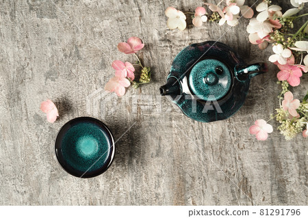Green tea in dark turquoise clay teapot and small cup on grey wooden background with pink and white flowers decoration. Creative minimalist tea ceremony concept. Copy space, top view. 81291796