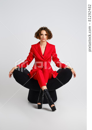Dancer woman in red pantsui  and pole dance boots sits in a soft chair with crossed legs 81292482