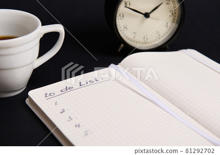 Cropped image of an opened organizer, notepad with list to do on blank white sheets, cup of coffee, alarm clock on black background with copy space. Business, Organization, Time Management concepts. 81292702