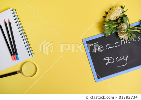 Flat lay of asters flowers lying down on chalkboard with text Teachers Day , and a magnifier loupe next to colored pencils on a blank sheets of a notepad isolated on yellow background with copy space 81292734