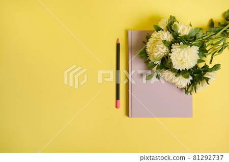 Flat lay. Beautiful delicate bouquet of Autumn flowers, white asters lying down on the corner of a notepad organizer, next to a wooden color pencil, isolated over yellow background with copy space 81292737
