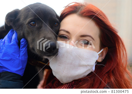 Young female using a face mask as a coronavirus spreading prevention walking with her dog. Global COVID-19 pandemic concept image. 81300475