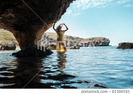 A woman in a white dress and a black hat standing on the rocks by the sea 81300591