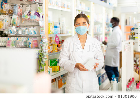 Portrait of a young female pharmacist in a protective mask demonstrating in her hands a goods Portrait of a young female pharmacist in a protective mask demonstrating in her hands a goods 81301906
