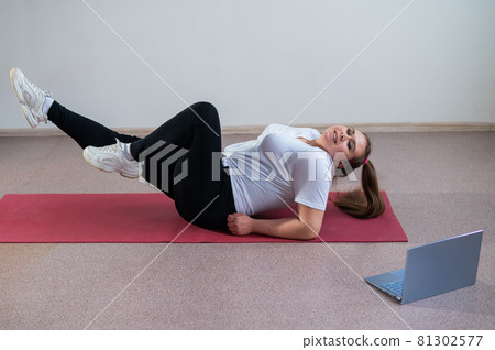 A chubby young woman is watching an online fitness lesson on a laptop. Distance sports training. A chubby young woman is watching an online fitness lesson on a laptop. Distance sports training. 81302577