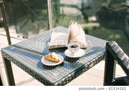 Cup of black coffee, freshly baked croissant and open book on wicker rattan table on hotel balcony 81314182