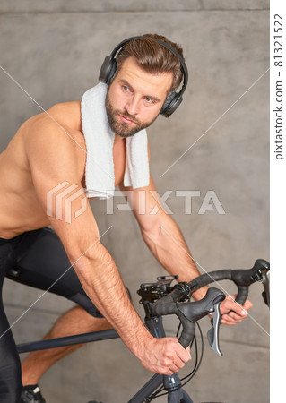 Bearded young man in headphones riding stationery bike Bearded young man in headphones riding stationery bike 81321522