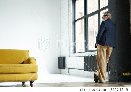 Full length shot of stylish middle aged man in business casual wear and sunglasses leaning on the wall, looking away while posing in loft interior 81321549