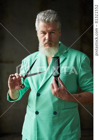 Portrait of gray haired mature man with beard wearing colorful outfit looking at camera, holding sharp barber scissors and hair comb, standing over dark background 81321552