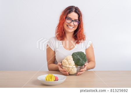 Caucasian woman prefers healthy food. Redhead girl chooses between broccoli and donuts on white background. Caucasian woman prefers healthy food. Redhead girl chooses between broccoli and donuts on white background. 81325692