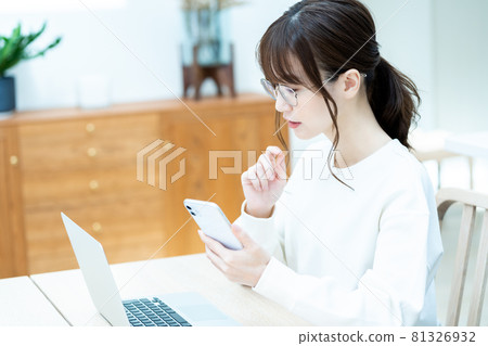 A woman with glasses who operates a smartphone while using a laptop in the living room 81326932