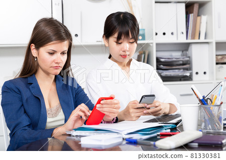 Two women write sms by phone in the office Two women write sms by phone in the office 81330381