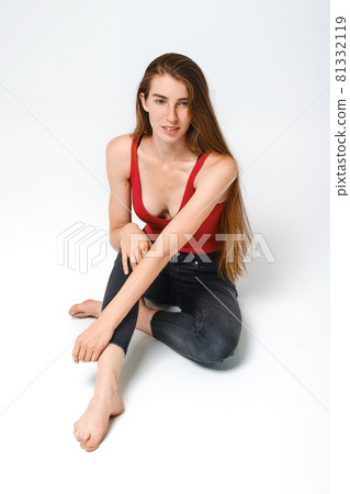 Cute young woman in red tank-top and grey jeans sits on the floor Cute young woman in red tank-top and grey jeans sits on the floor 81332119