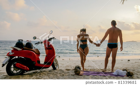 Scooter road trip. Lovely couple on red motorbike in white clothes on sand beach. People walking near the tropical palm trees, sea. Motorcycle rent. Scooter road trip. Lovely couple on red motorbike in white clothes on sand beach. People walking near the tropical palm trees, sea. Motorcycle rent. 81339681