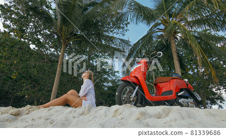 Scooter road trip. Woman alone on red motorbike in white clothes on sand beach by ocean. One girl caucasian tourist walk near tropical palm tree. 81339686