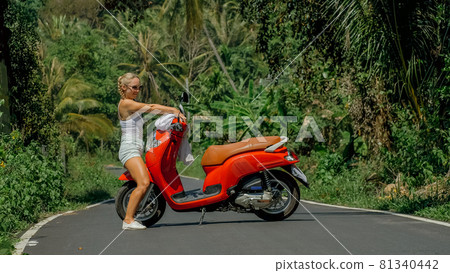 Woman on red scooter in white clothes drive on forest road trail. Dancing trip. One girl caucasian tourist in sunglasses dance, relax near motorbike. Woman on red scooter in white clothes drive on forest road trail. Dancing trip. One girl caucasian tourist in sunglasses dance, relax near motorbike. 81340442