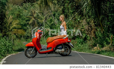 Woman on red scooter in white clothes drive on forest road trail. Dancing trip. One girl caucasian tourist in sunglasses dance, relax near motorbike. 81340443
