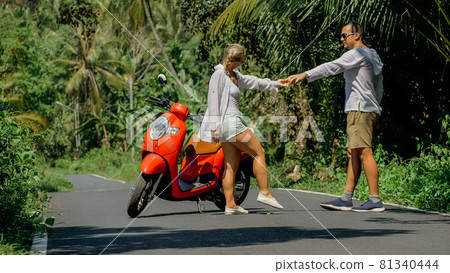 Love couple on red motorbike in white clothes, sunglasses on forest road trail trip. Dancing road. Two caucasian tourist woman man dance near scooter. Love couple on red motorbike in white clothes, sunglasses on forest road trail trip. Dancing road. Two caucasian tourist woman man dance near scooter. 81340444