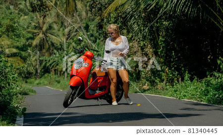 Woman on red scooter in white clothes drive on forest road trail. Dancing trip. One girl caucasian tourist in sunglasses dance, relax near motorbike. Woman on red scooter in white clothes drive on forest road trail. Dancing trip. One girl caucasian tourist in sunglasses dance, relax near motorbike. 81340445