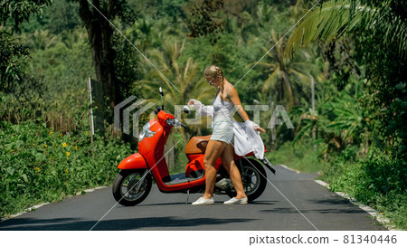 Woman on red scooter in white clothes drive on forest road trail. Dancing trip. One girl caucasian tourist in sunglasses dance, relax near motorbike. Woman on red scooter in white clothes drive on forest road trail. Dancing trip. One girl caucasian tourist in sunglasses dance, relax near motorbike. 81340446