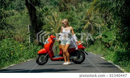Woman on red scooter in white clothes drive on forest road trail. Dancing trip. One girl caucasian tourist in sunglasses dance, relax near motorbike. Woman on red scooter in white clothes drive on forest road trail. Dancing trip. One girl caucasian tourist in sunglasses dance, relax near motorbike. 81340447