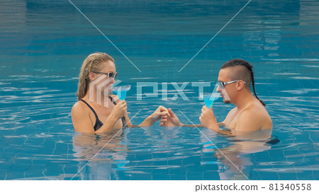 The loving couple hugs and kisses, drinking blue cocktail alcohol liquor in swimming pool at hotel outdoor. Portrait of caucasian man and woman. The loving couple hugs and kisses, drinking blue cocktail alcohol liquor in swimming pool at hotel outdoor. Portrait of caucasian man and woman. 81340558